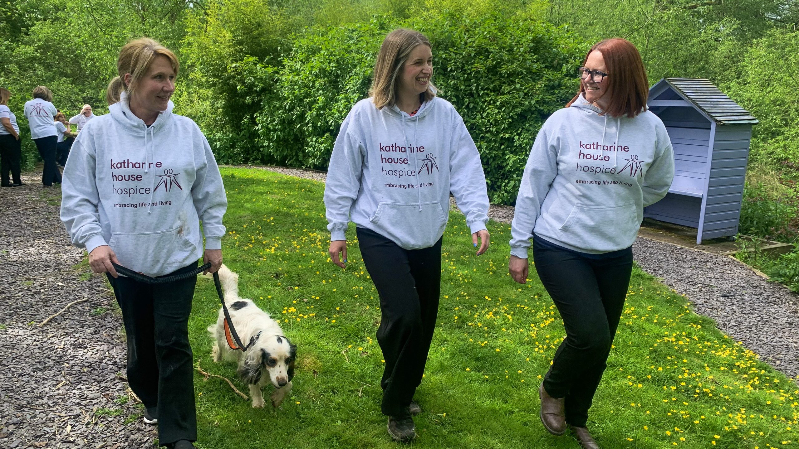 Group of three women walking, talking and smiling – all wearing Katharine House Hospice hoodies. Left most woman is walking a spaniel.