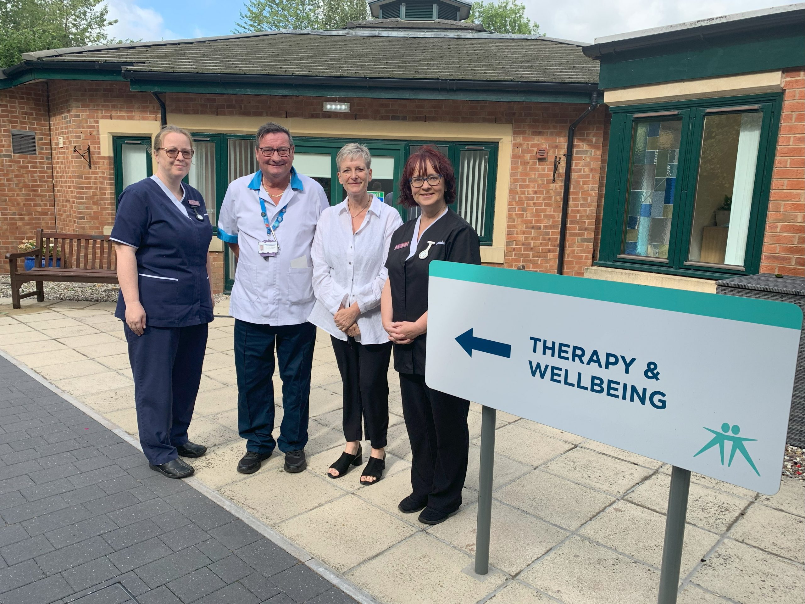 Left to right: Deputy Outpatient Lead Nicola, Therapy Practitioner Iain, Amanda Brooke and Complementary Therapist Rachel. All standing outside next to the 'Therapy & wellbeing' sign.