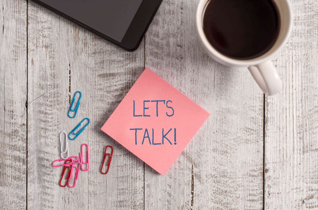 Picture showing a coffee cup with black coffee and a pink note that says LET'S TALK on a wooden table top