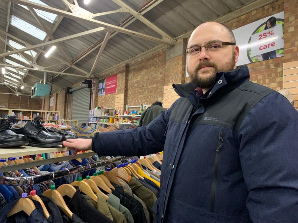 A man with a beard and glasses standing next to the menswear rail inside the shop